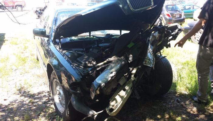Severely damaged dark sedan with a crushed front end and popped hood after a car crash.
