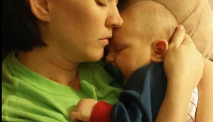 A mother cradles her sleeping baby, resting her forehead gently against the infant's head.
