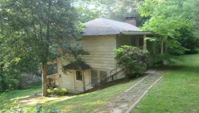 A rustic two-story wooden house nestled on a grassy hillside surrounded by dense green trees.