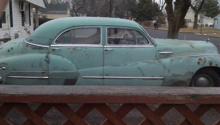 Side profile of a rusty, light green vintage 1940s sedan parked in front of a suburban house.