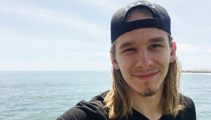 A smiling young man with long blonde hair and a backward cap, with the ocean stretching out behind him.