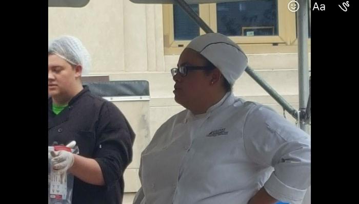 A chef in a white coat and cap standing outdoors next to a culinary worker in a hairnet.