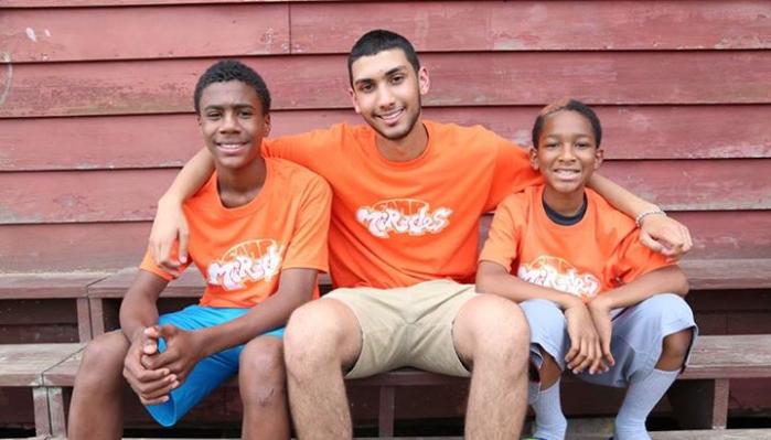 Three smiling young men in matching orange camp t-shirts sitting together on wooden bleachers.