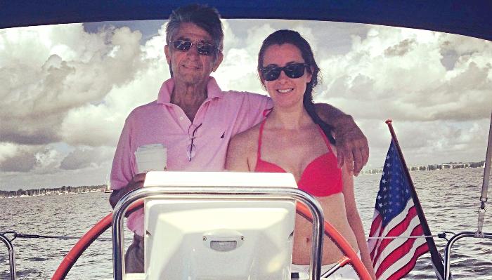 A man in a pink polo and a woman in a red bikini smile at the helm of a boat with an American flag behind them.