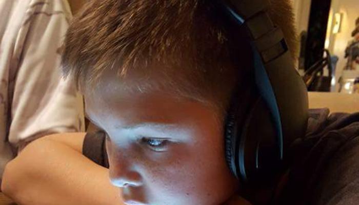 A young boy wearing black over-ear headphones looks intently at a glowing screen.