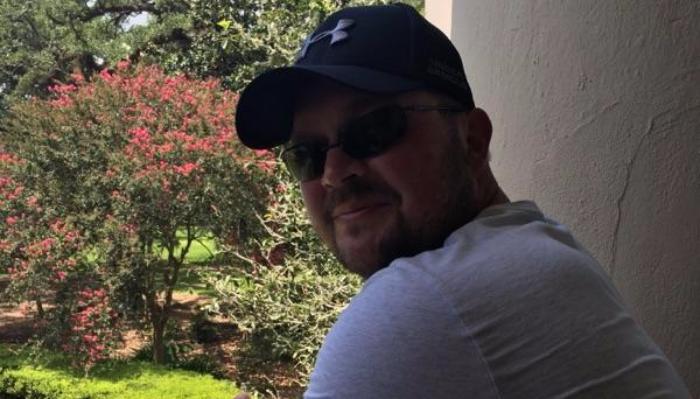 Smiling man with a beard wearing a black cap, sunglasses, and white t-shirt outdoors near a flowering pink tree.