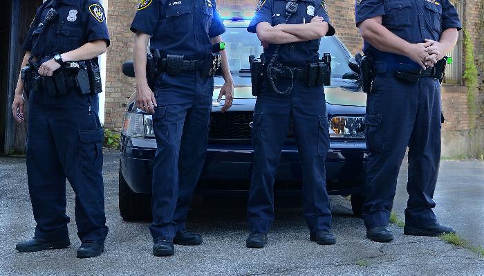 Four police officers in blue uniforms stand side-by-side in front of a police cruiser.