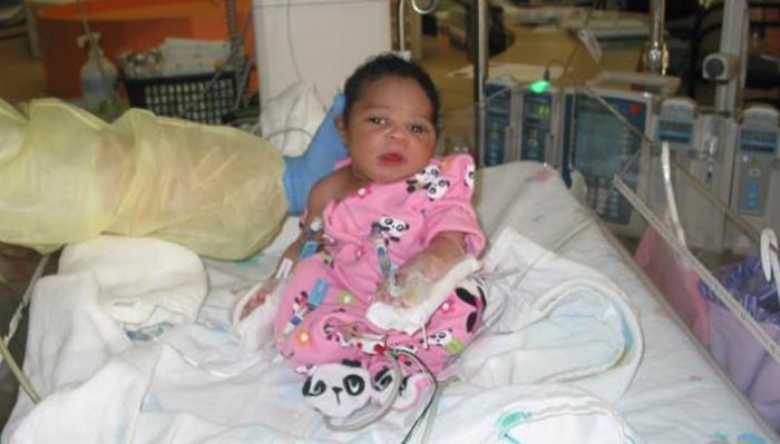 A baby wearing pink panda pajamas sits in a hospital crib surrounded by medical equipment.