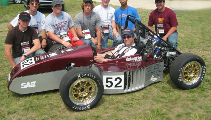 A student engineering team poses with a driver in their maroon and silver number 52 Formula SAE race car.
