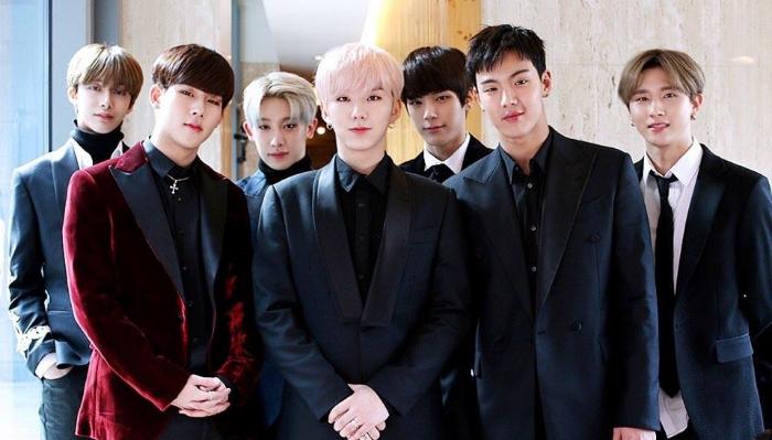 Seven members of K-pop group Monsta X pose together indoors wearing stylish dark suits and tuxedos.