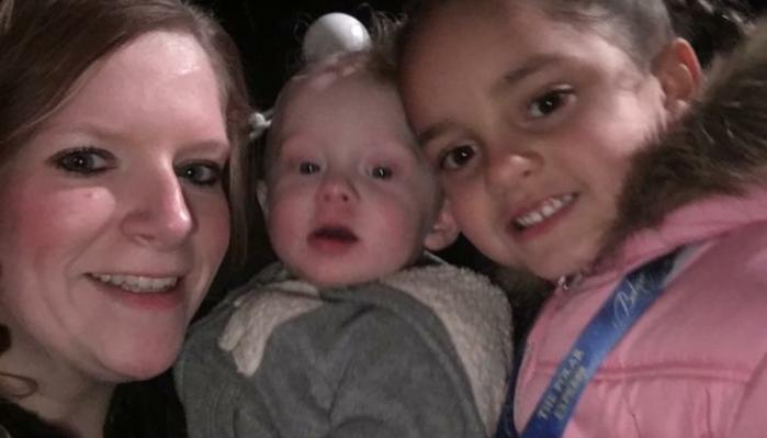 A smiling mother takes a close-up selfie with her baby and young daughter wearing a pink coat.
