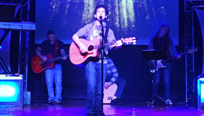 A young musician sings and plays acoustic guitar on stage with a live band under blue lighting.