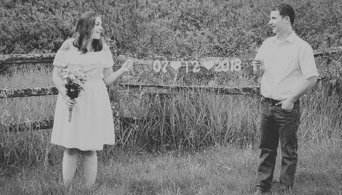 A couple standing in a grassy field holds a banner reading 07-12-2018 in front of a wooden fence.