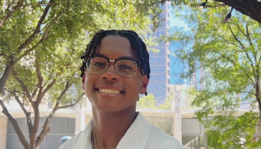 A smiling young Black man wearing glasses, standing outdoors in front of leafy green trees and city buildings.