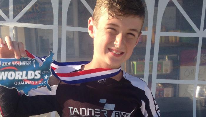 A smiling boy wearing a BMX jersey holds up a UCI USA BMX Worlds Qualified Rider medal.