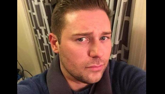 Close-up portrait of a man with short brown hair, blue eyes, and light stubble wearing a blue sweater.