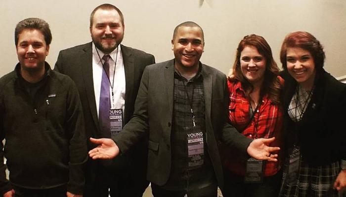 Five smiling young adults pose together at a conference while wearing lanyard badges.