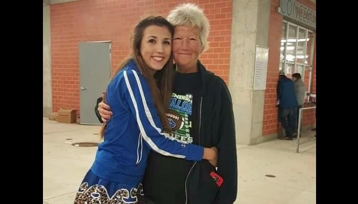 A smiling young woman in a blue jacket hugs an older woman with short grey hair near a brick building.
