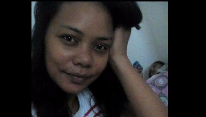 Close-up selfie of a woman resting her head on her hand, with a child lying in bed in the background.