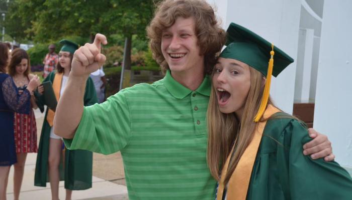 A smiling young man poses excitedly with a female graduate wearing a green cap, gown, and gold sash.