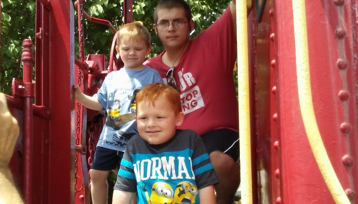 A young man and two young boys smiling as they play together on a bright red playground structure.