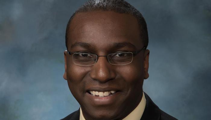 Smiling headshot of a Black man wearing rectangular glasses against a blue studio background.