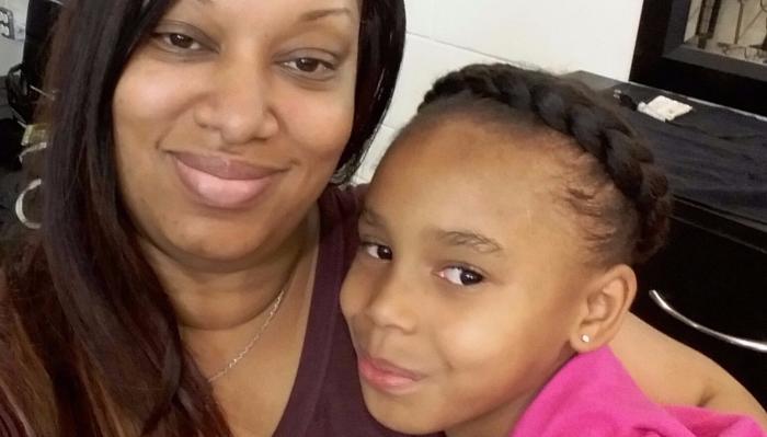 A smiling mother poses close to her young daughter, who wears a braided crown hairstyle.