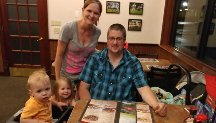 A smiling family of four with two young children poses at a restaurant table.