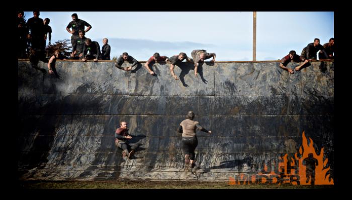 Participants atop a steep muddy wall reach down to help a runner climb up during an obstacle course race.