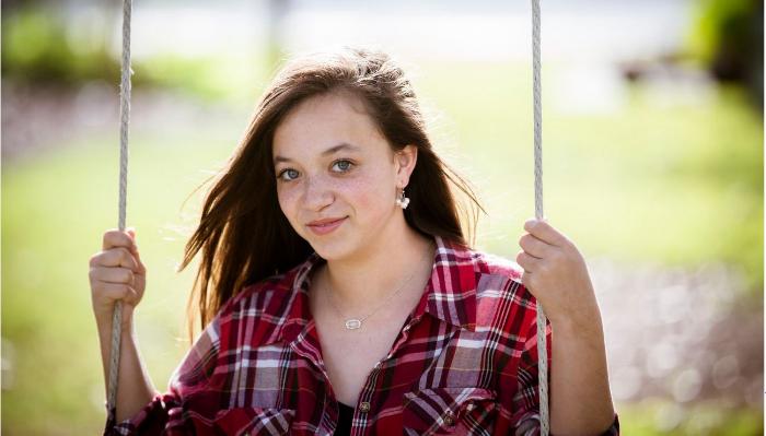 A young woman wearing a red plaid shirt sits on a swing outdoors, smiling gently at the camera.