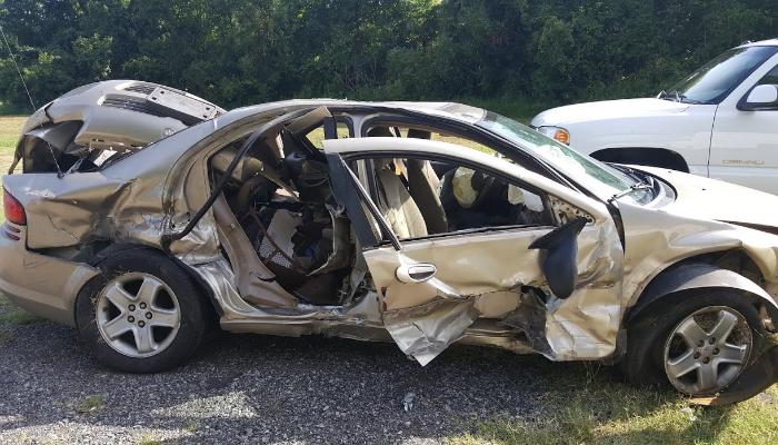 Severely wrecked gold sedan with extensive side impact damage, crushed roof, and deployed airbags.