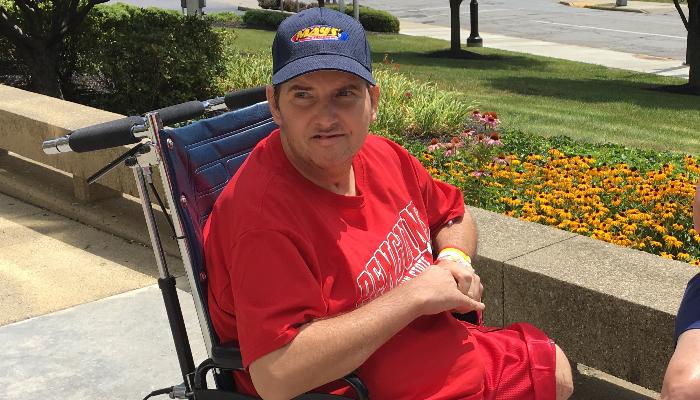 A man in a red shirt and blue baseball cap sitting in a wheelchair outdoors near a flower garden.