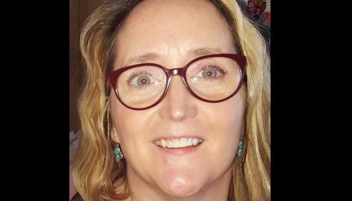 Close-up of a smiling woman with wavy blonde hair, red glasses, and turquoise earrings.