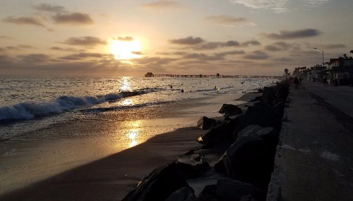 Golden sunset reflecting over ocean waves on a sandy beach with rocks and a long wooden pier in the distance.