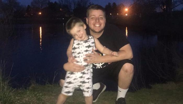 A smiling man hugs a happy young boy outdoors at night near a lake with city lights reflecting on the water.