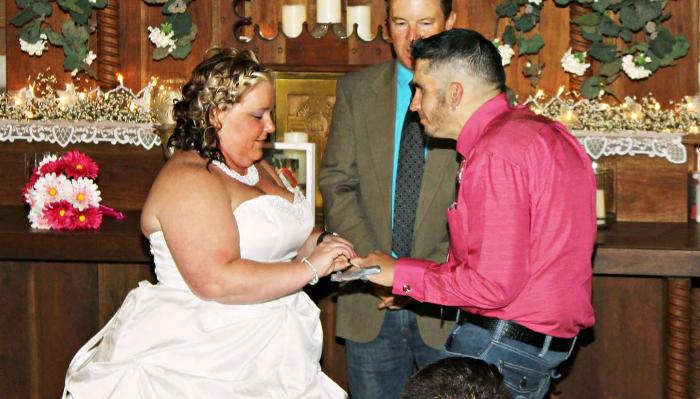 A bride in a white gown and a groom in a bright pink shirt exchange wedding rings at the altar.