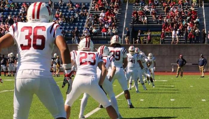 Football players in white and red uniforms lined up on a field in front of crowded stadium bleachers.