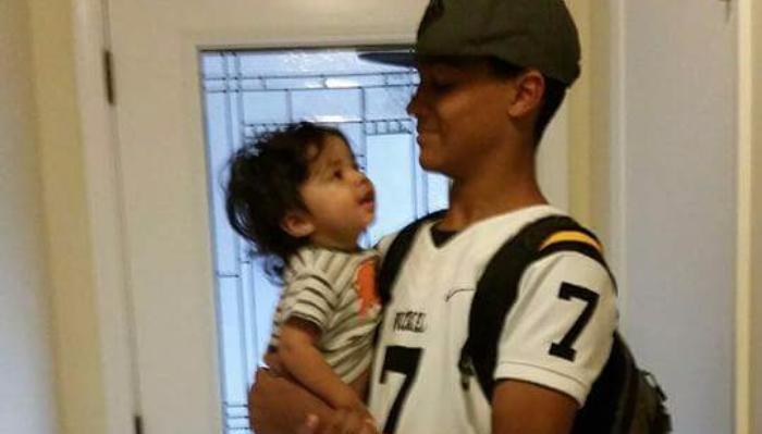 A teenage boy wearing a sports jersey holds a toddler in his arms while looking at them affectionately.
