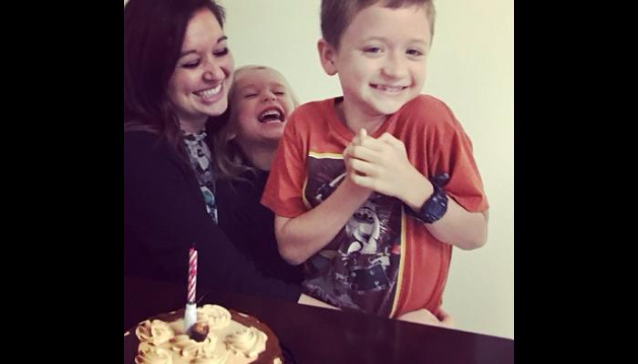 A happy mother and two laughing children gather behind a frosted birthday cake with a single candle.