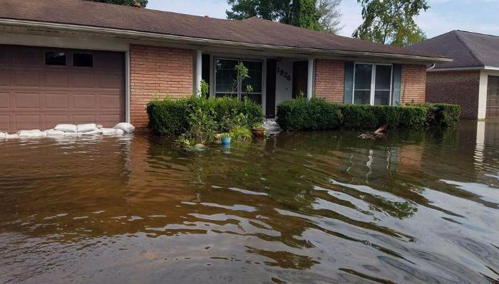 Deep floodwater surrounds a single-story brick home, with sandbags stacked in front of the garage door.