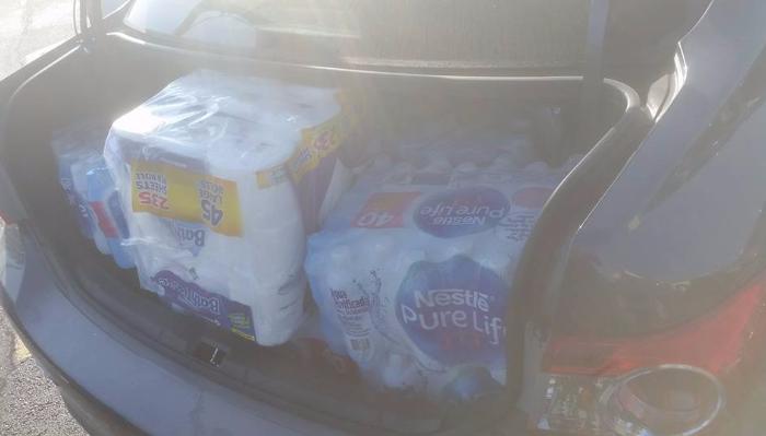 An open car trunk packed with cases of bottled water and bulk toilet paper.