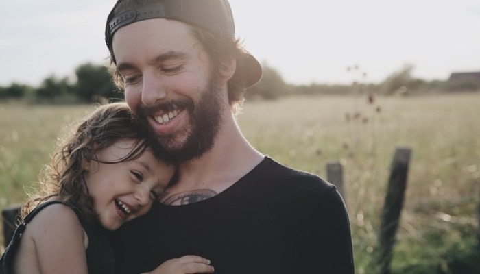 A smiling bearded man in a backwards cap holds a laughing young child resting her head on his chest.