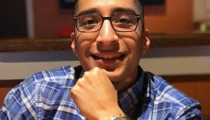 Smiling young man wearing glasses and a blue plaid shirt, resting his chin on his hand.