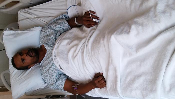 A man lying in a hospital bed wearing a patterned hospital gown and covered by a white blanket.