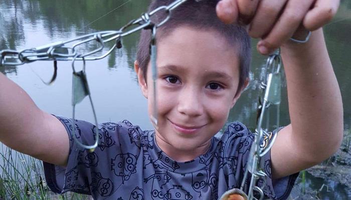 A smiling young boy holds up a metal fish stringer by a lake.