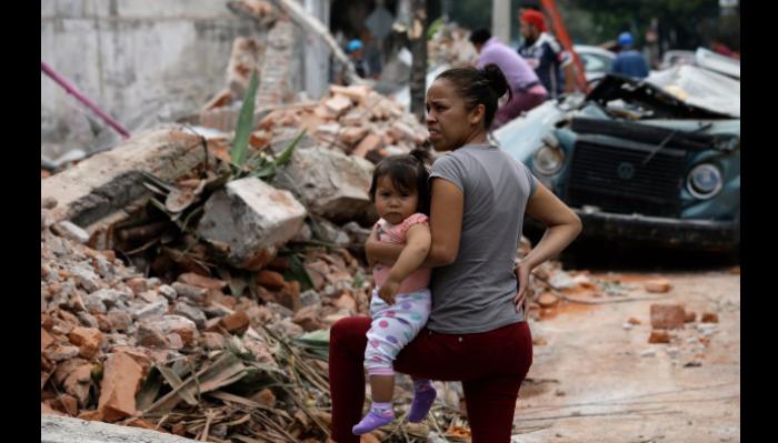 A worried mother holds her young child amidst piles of rubble and a crushed car after a disaster.