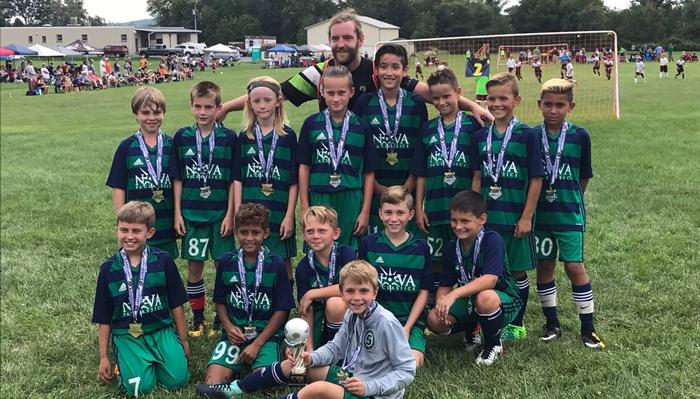 A youth soccer team wearing green jerseys and medals poses with their coach and a trophy on a grass field.