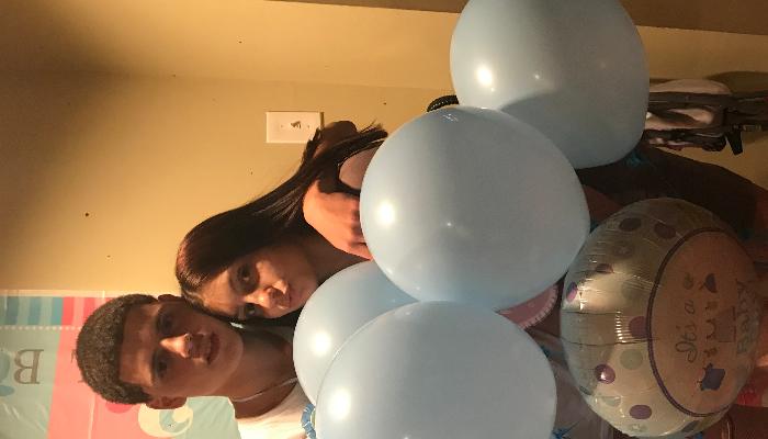 A smiling young couple posing behind light blue balloons and an "It's a Boy" foil balloon.