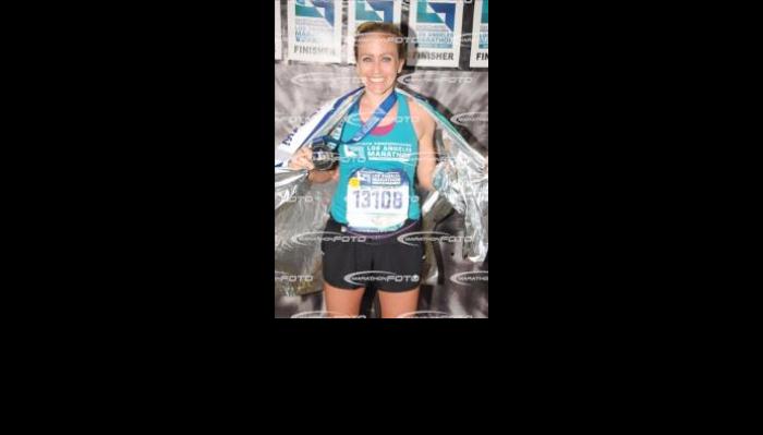 A smiling woman wearing a medal, race bib, and thermal blanket after completing the Los Angeles Marathon.