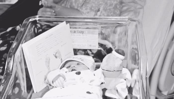 Newborn baby lying in a clear hospital bassinet next to a card with footprints and an "It's a Boy" label.